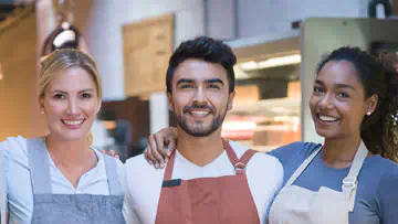 Smiling people with aprons stand in front of a cafe counter.